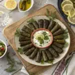 Greek Dolmades Guide — platter of homemade stuffed grape leaves with lemon, dips, and Greek salad